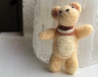 Pode incluir: Um pequeno urso de peluche de feltro bege com uma echarpe listrada marrom e branca, em pé sobre uma superfície branca.