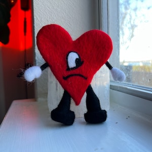 Op de afbeelding: Een rood vilt hart met een verdrietig gezicht en één groot oog, met een zwarte broek en witte handschoenen. Het hart zit op een witte vensterbank.