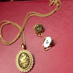 May include: A gold chain necklace with a black and gold oval pendant featuring a bird design. The pendant is surrounded by small clear stones. The necklace comes with a pair of matching earrings with black and gold designs.