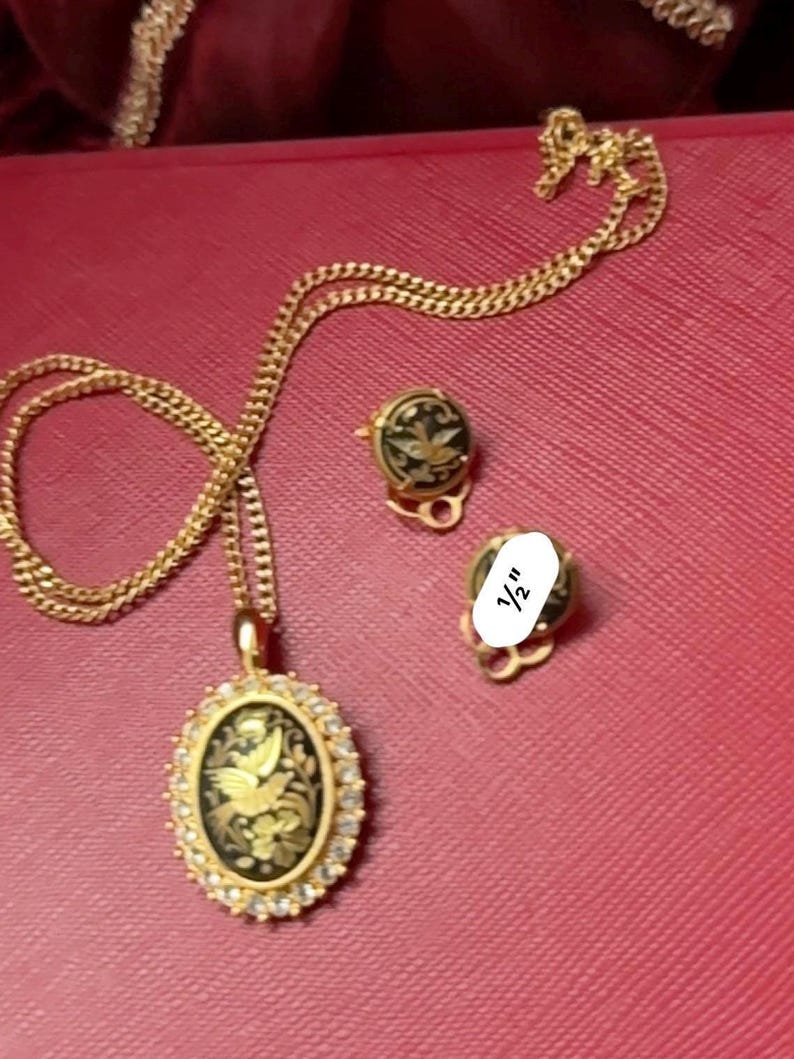 May include: A gold chain necklace with a black and gold oval pendant featuring a bird design. The pendant is surrounded by small clear stones. The necklace comes with a pair of matching earrings with black and gold designs.