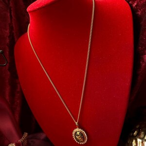 May include: A gold chain necklace with a gold oval pendant featuring a black and gold design. The necklace is displayed on a red velvet mannequin.
