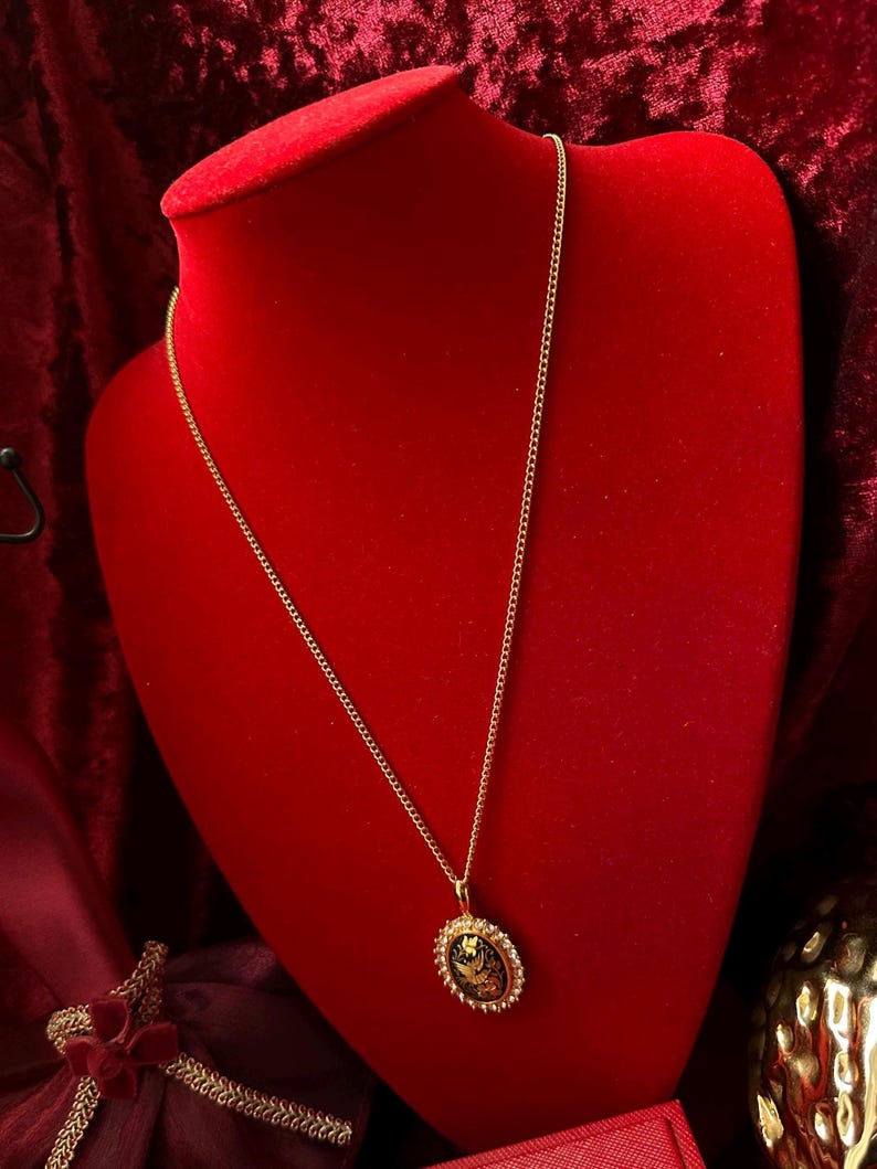 May include: A gold chain necklace with a gold oval pendant featuring a black and gold design. The necklace is displayed on a red velvet mannequin.