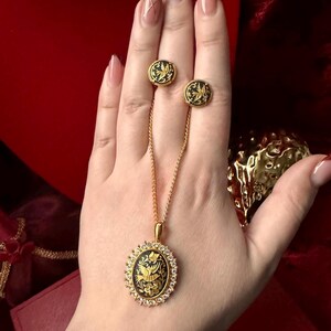 May include: A gold necklace with a black and gold oval pendant featuring a bird design. The pendant is surrounded by small clear stones. The necklace is paired with matching earrings.