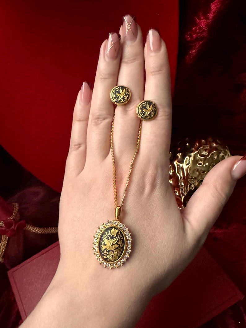 May include: A gold necklace with a black and gold oval pendant featuring a bird design. The pendant is surrounded by small clear stones. The necklace is paired with matching earrings.