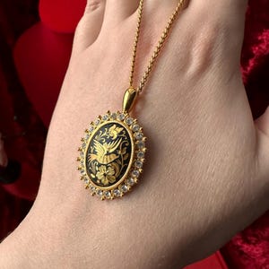May include: A gold necklace with a black oval pendant featuring a detailed gold bird design surrounded by clear rhinestones.