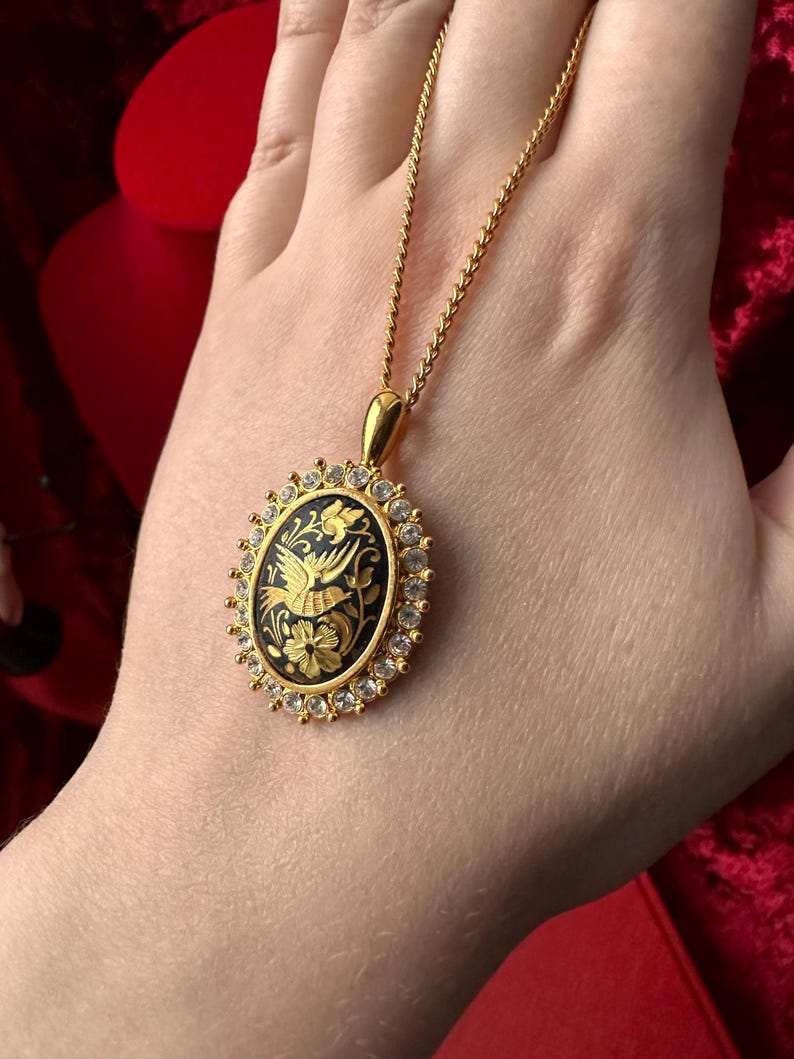 May include: A gold necklace with a black oval pendant featuring a detailed gold bird design surrounded by clear rhinestones.