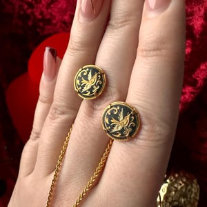 May include: A pair of gold earrings with black enamel and a gold bird design. The earrings are on a hand wearing a gold chain necklace.