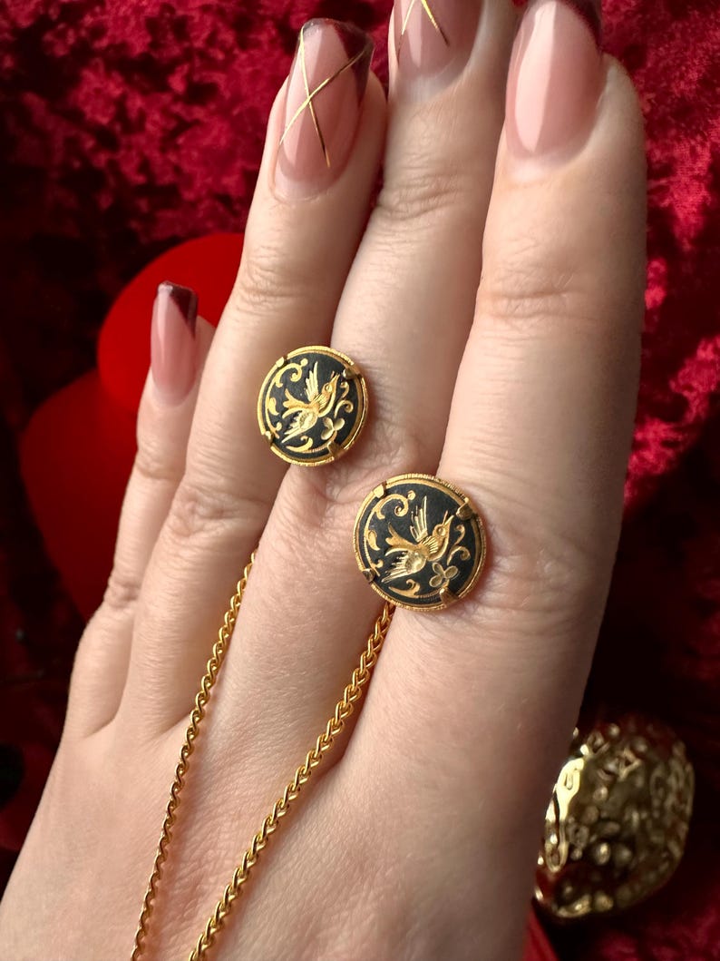 May include: A pair of gold earrings with black enamel and a gold bird design. The earrings are on a hand wearing a gold chain necklace.