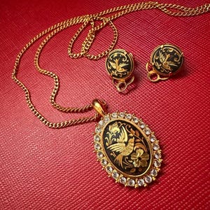 May include: A gold chain necklace with a black and gold oval pendant featuring a bird design. The pendant is surrounded by clear rhinestones. The necklace comes with a pair of matching earrings with black and gold bird designs.