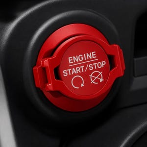 May include: A circular red engine start/stop button cover featuring a flip-up lid design. The surface is printed with the white text "ENGINE START/STOP" above two circular arrow icons, one indicating a clockwise rotation and the other a crossed-out symbol.