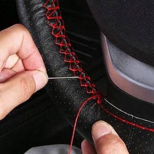 May include: A black perforated steering wheel cover featuring a cross-stitch pattern created with bright red thread. The cover is being installed onto a steering wheel using a silver needle and red thread to secure the edges together.