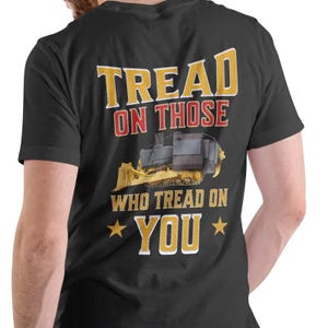 May include: Black t-shirt with the text "TREAD ON THOSE WHO TREAD ON YOU" in yellow and red lettering. A yellow and gray bulldozer graphic is centered below the text. Two yellow stars flank the word "YOU".