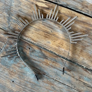 May include: A silver metal headband with a sunburst design. The headband is made of thin metal strips that radiate out from the center.
