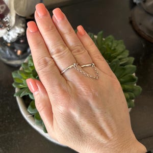 May include: A silver ring with a delicate chain connecting two bands. The ring is worn on the index and middle finger.