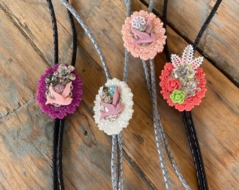 May include: Four bolo ties with different floral and bird designs. The bolo ties are made with silver and black cords and feature pink, white, and purple flowers with silver accents.