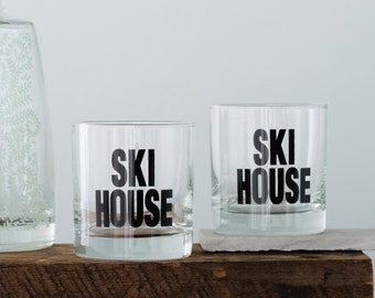 May include: Two clear glass tumblers with black text that reads "SKI HOUSE". The glasses are sitting on a wooden surface.