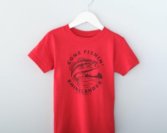 May include: A bright red short-sleeved t-shirt featuring a black graphic print on the chest. The design depicts a fish jumping out of water with the text GONE FISHIN' RHINELANDER WISCONSIN arranged in a circular pattern around the illustration.
