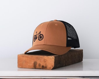 May include: A brown baseball cap with a black mesh back and a curved brim. The cap features a black embroidered bicycle design on the front. The hat is resting on a wooden block.