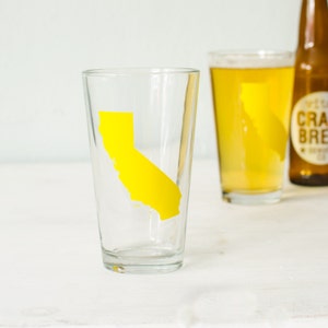 May include: A clear glass pint glass with a yellow outline of the state of California on the side. The glass is sitting on a white surface.