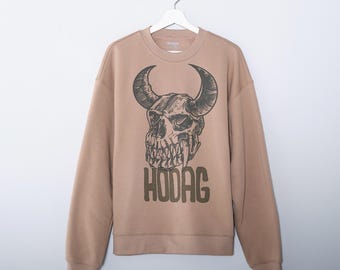 May include: Tan crewneck sweatshirt featuring a detailed skull graphic with large horns and the word "HODAG" printed below. The design is in a dark brown color. The sweatshirt is hanging on a white hanger.