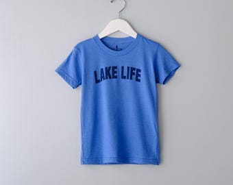 May include: A short-sleeved blue t-shirt featuring the text LAKE LIFE printed in dark blue across the chest. The garment is displayed on a white plastic hanger, showcasing a crew neckline and a standard fit with a simple, clean design.