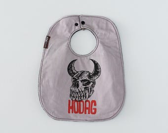 May include: A light gray baby bib featuring a black ink illustration of a horned skull with sharp teeth. Below the skull, the word HODAG is printed in bold red capital letters. The bib has black trim and two black snap closures at the neck.