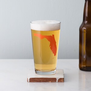 May include: A pint glass with a frosted rim and a clear body filled with a light-colored beer. The glass has an orange outline of the state of Florida on the side.