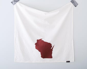 May include: White kitchen towel with a maroon outline of the state of Wisconsin. The towel is hanging on a light gray wall, secured with gray tape at the top corners. A small black tag is visible at the bottom right corner.