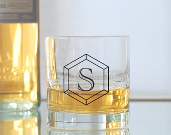 May include: A clear glass tumbler with a black geometric design featuring the letter "S" in the center. The glass is filled with a golden liquid.