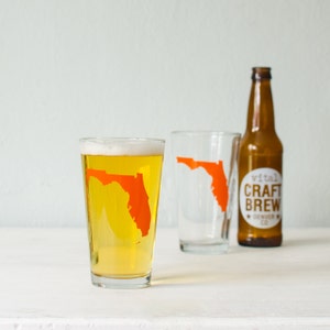 May include: Two clear glass pint glasses with an orange outline of the state of Florida. One glass is filled with a light yellow beer. A brown glass bottle with a white label that reads "Vital Craft Brew Denver Co." is in the background.