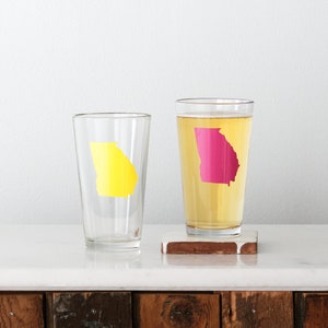 May include: Two clear glass tumblers with a state of Georgia outline design. One tumbler has a yellow outline and the other has a pink outline.