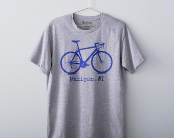 May include: A heather gray short-sleeve t-shirt featuring a blue graphic of a road bicycle centered on the chest. Below the bicycle graphic, the text reads Madison, WI in a blue typewriter-style font. The shirt has a crew neckline and a medium size label.