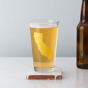 May include: A clear glass pint glass with a gold outline of the state of California. The glass is filled with a light yellow beer.