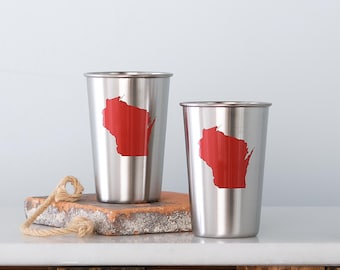 May include: Two stainless steel tumblers with a red Wisconsin state outline design. The tumblers are cylindrical with a rolled rim. They are displayed on a brick-like surface with a rope detail, against a white background.
