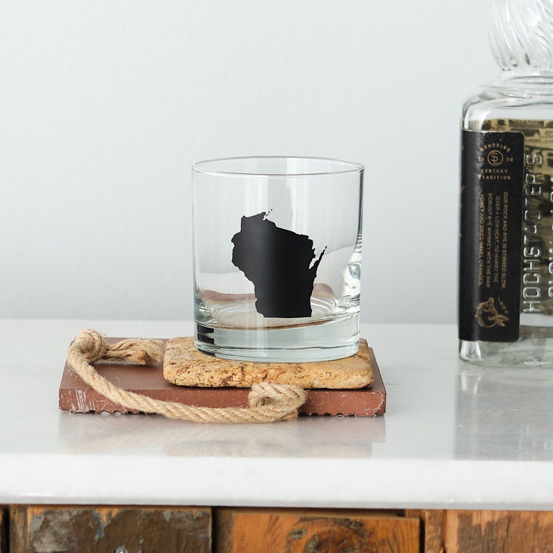 May include: A clear glass rocks glass with a black silhouette of the state of Wisconsin on the side. The glass is sitting on a brown cork coaster.