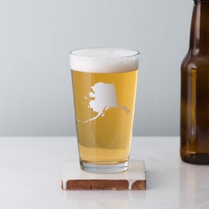 May include: A clear glass pint glass with a white outline of the state of Alaska on the side. The glass is filled with a light-colored beer and has a white foam head.