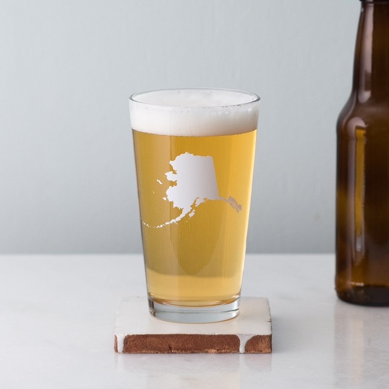 May include: A clear glass pint glass with a white outline of the state of Alaska on the side. The glass is filled with a light-colored beer and has a white foam head.