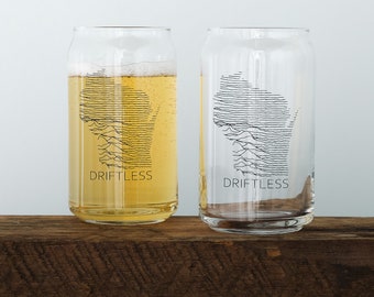 May include: Two clear glass drinking glasses with a black and white outline of the state of Wisconsin. The glasses have the word "Driftless" printed on the bottom.