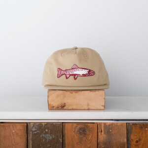 Puede incluir: Gorra de béisbol beige con un diseño de pez granate. La gorra tiene una visera plana y un diseño clásico de seis paneles. La ilustración del pez es detallada, con escamas y aletas visibles. El sombrero se exhibe sobre un bloque de madera.