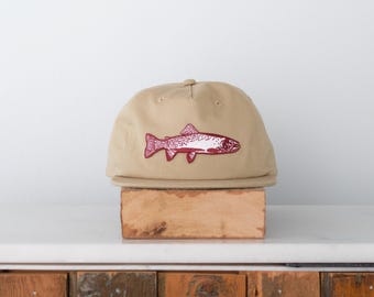 May include: A tan baseball cap featuring a maroon fish graphic. The cap has a flat brim and a classic six-panel design. The fish illustration is detailed, with visible scales and fins. The hat is displayed on a wooden block, suggesting a rustic or outdoorsy theme.