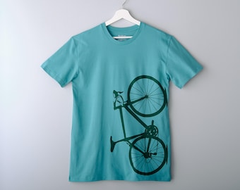 May include: A turquoise t-shirt with a dark green bicycle graphic. The bicycle design is a stylized silhouette, featuring the wheels, frame, and handlebars. The t-shirt is hanging on a white hanger against a light gray background.