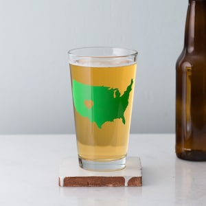May include: A clear glass pint glass with a green outline of the United States with a heart in the middle. The glass is filled with a light yellow beer.