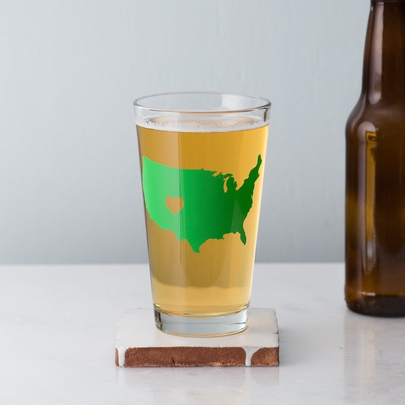 May include: A clear glass pint glass with a green outline of the United States with a heart in the middle. The glass is filled with a light yellow beer.