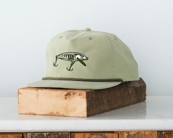 May include: A light green baseball cap with a green and white fish lure graphic. The cap has a rope band around the crown.