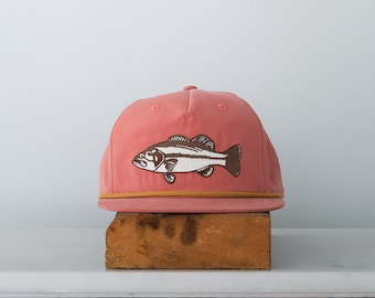May include: A coral pink baseball cap with a brown and white embroidered bass fish design.