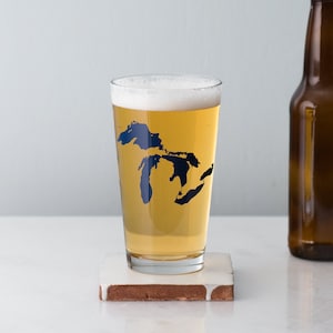 May include: A clear glass beer mug with a blue outline of the Great Lakes on the side. The mug is filled with a light-colored beer and is sitting on a brown coaster.