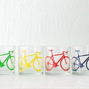 K&ouml;nnte beinhalten: Vier klare Glasbecher mit einem aufgedruckten Fahrradmotiv auf der Vorderseite. Die Fahrr&auml;der sind in Gr&uuml;n, Gelb, Rot und Blau.