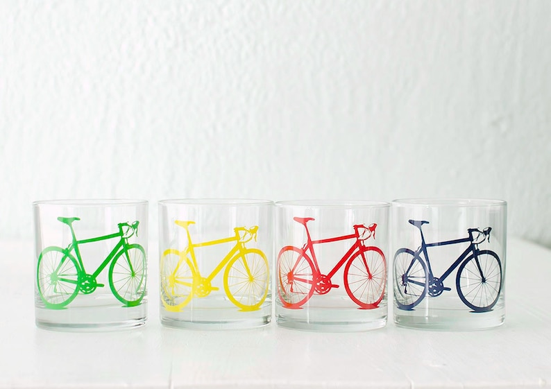 K&ouml;nnte beinhalten: Vier klare Glasbecher mit einem aufgedruckten Fahrradmotiv auf der Vorderseite. Die Fahrr&auml;der sind in Gr&uuml;n, Gelb, Rot und Blau.
