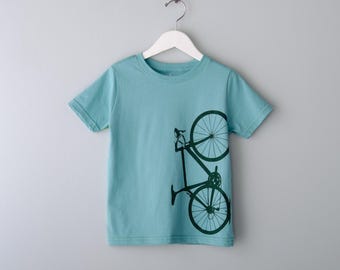 May include: A light blue t-shirt featuring a dark green bicycle graphic. The bicycle design is printed vertically on the front of the shirt. The shirt is hanging on a white hanger against a light gray background.