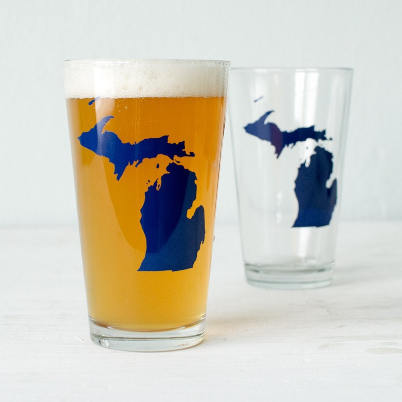 May include: Two clear glass pint glasses with a blue outline of the state of Michigan. One glass is filled with a light yellow beer.